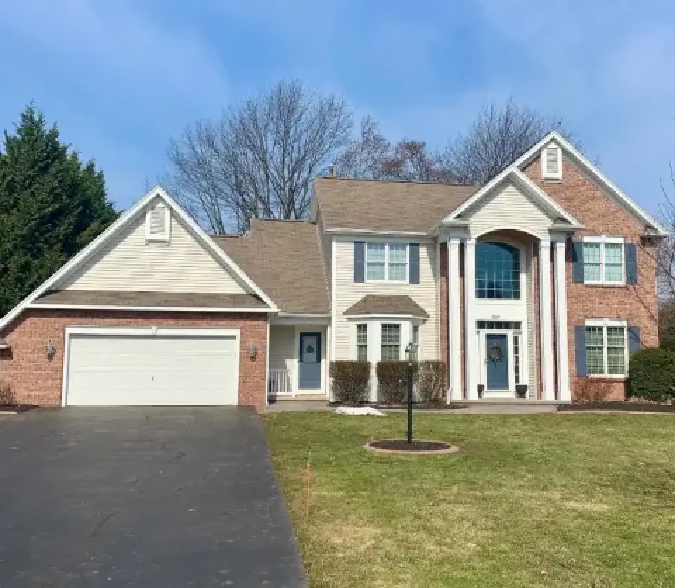Two-story suburban brick house with columns, attached garage, and a well-kept front lawn.