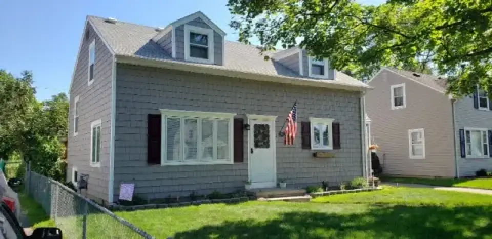 Gray two-story house with dormer windows, an American flag, and a small green lawn.