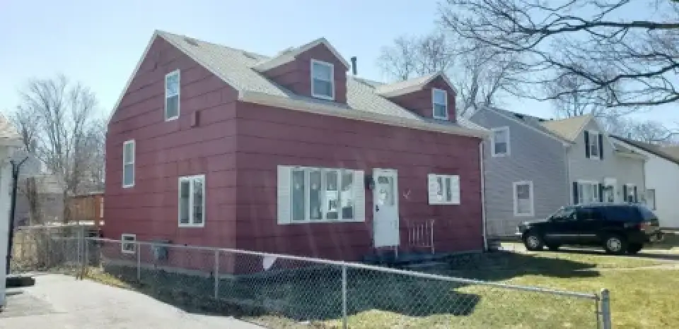 A red two-story house with white trim, fenced yard, and a black SUV parked next door.