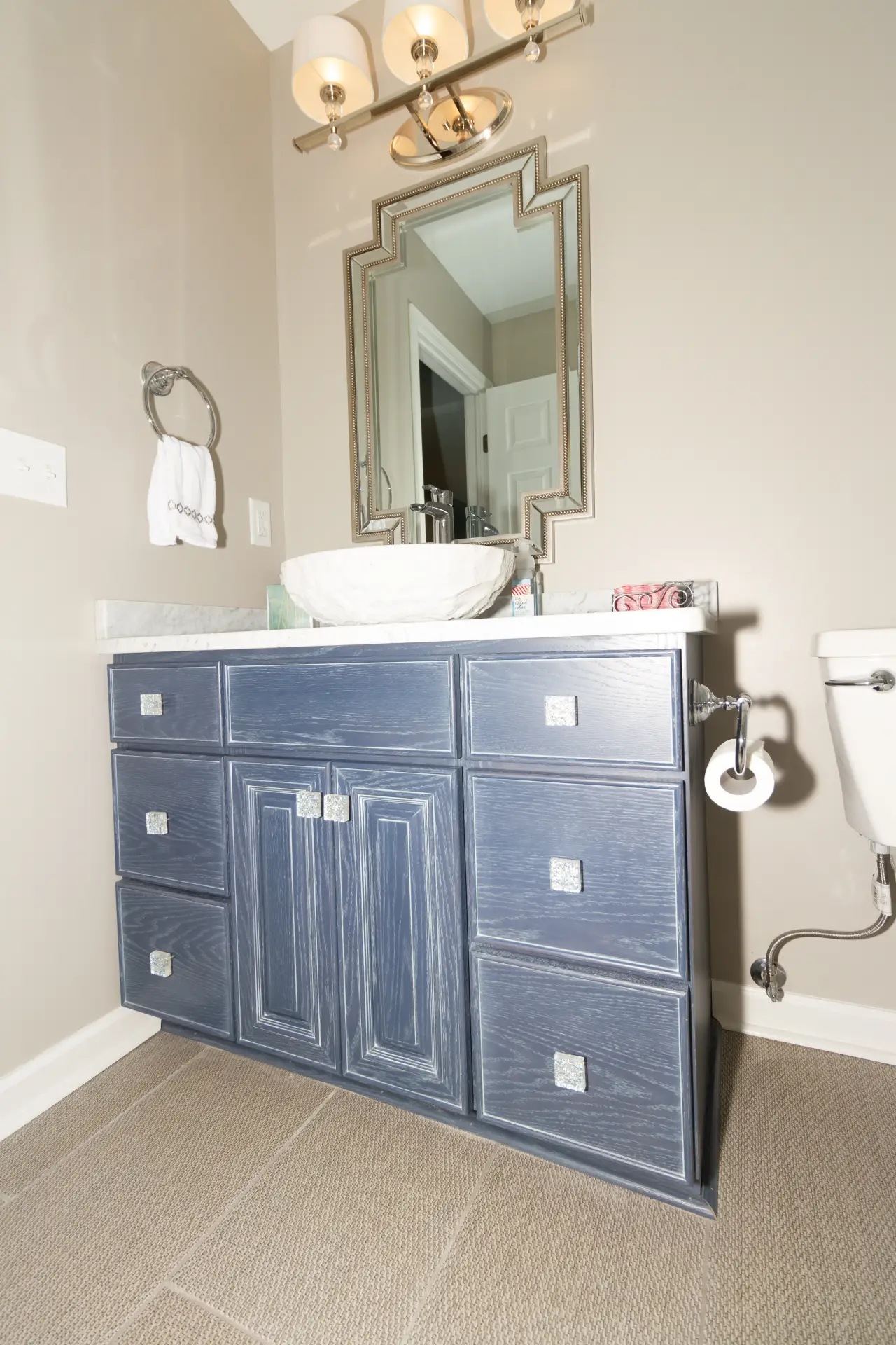 A modern bathroom featuring a blue vanity with multiple drawers, a round sink, a decorative mirror, and wall-mounted lighting.