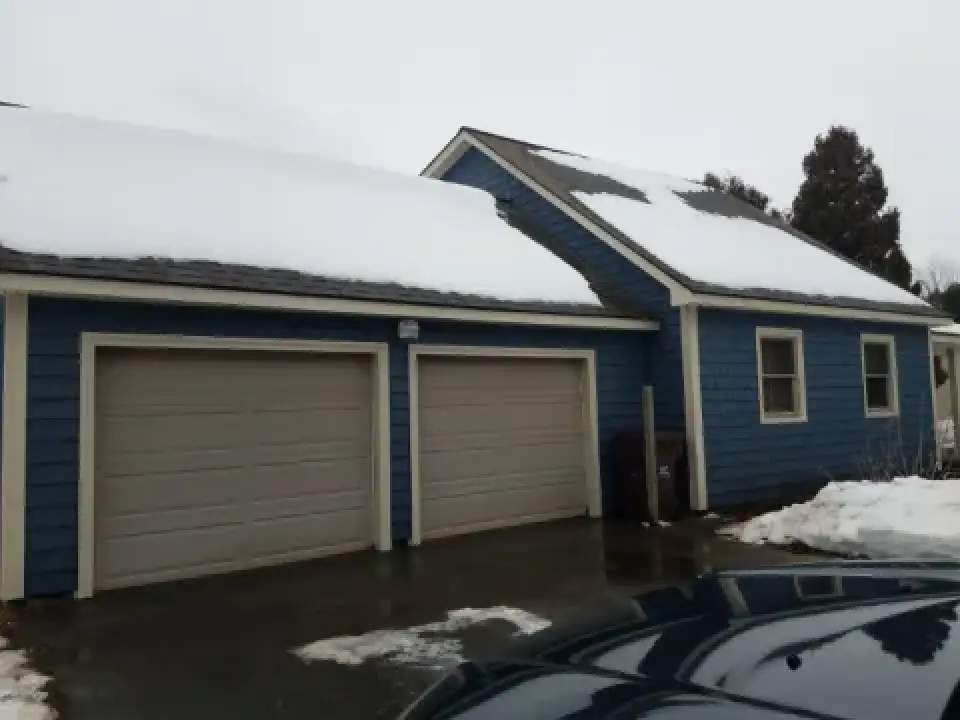 Blue house with a two-car garage, snow on the roof, and patches of snow on the ground.