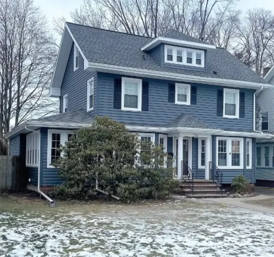 Two-story blue house with white trim, surrounded by bare trees and patches of snow on the lawn.