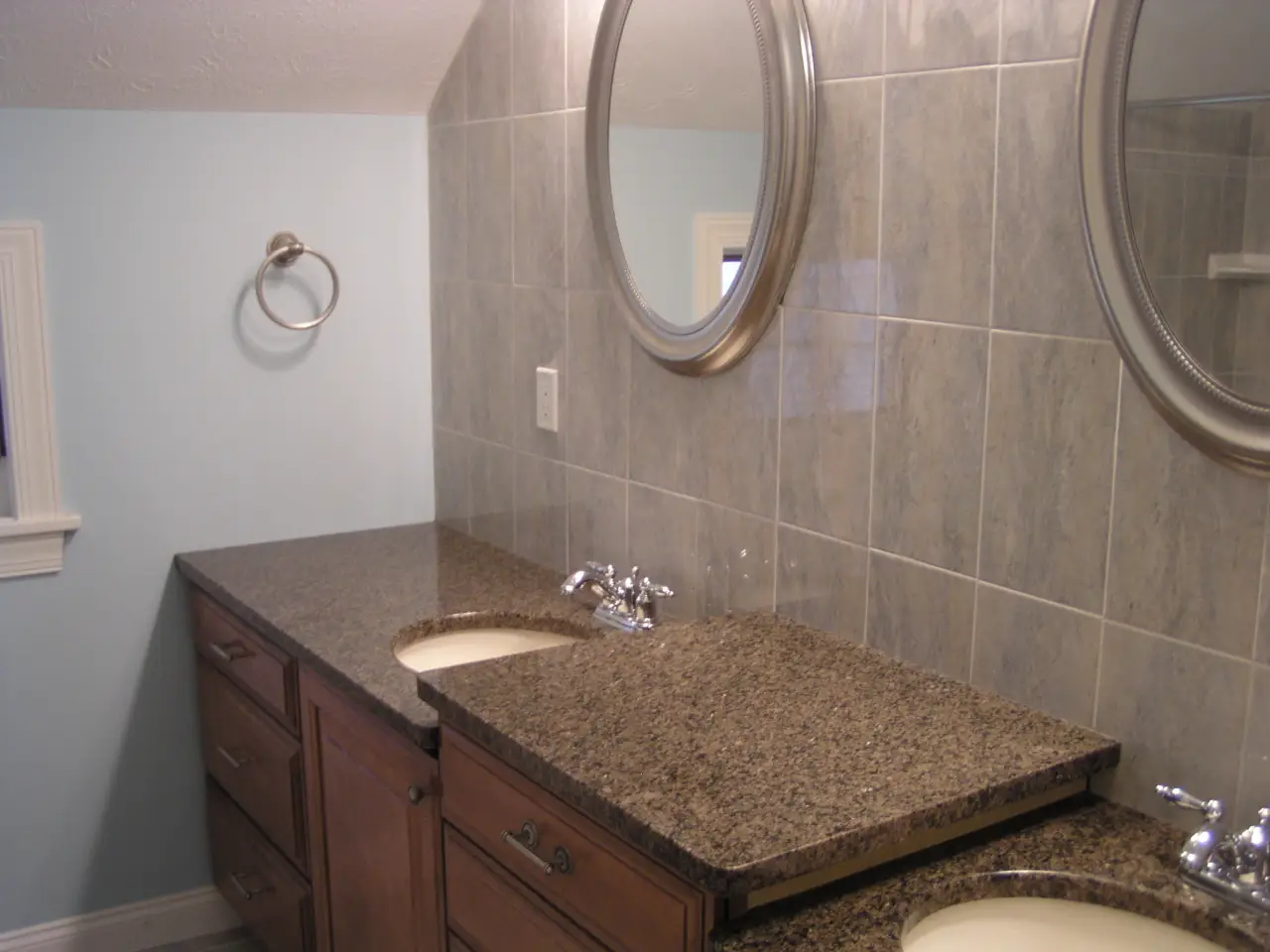 A modern bathroom with dual sinks, granite countertops, and round mirrors. Light blue walls complement the tiled backsplash.