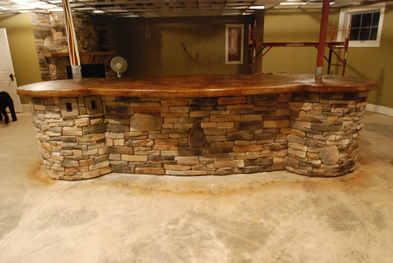 A curved stone bar with a wooden countertop in a spacious, unfinished room.