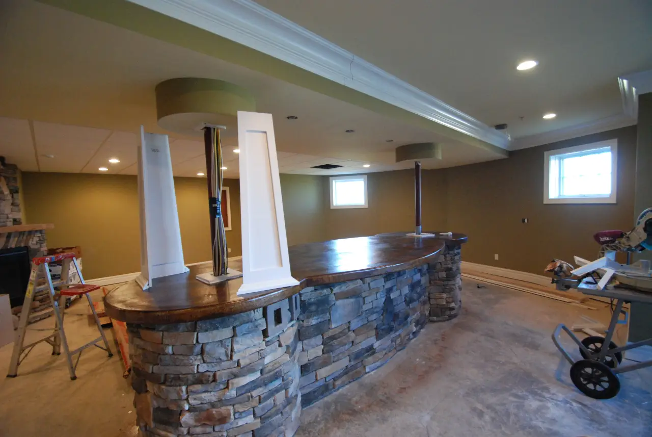 A partially finished basement with a stone bar, wooden countertop, and green walls. Tools and equipment are visible in the background.
