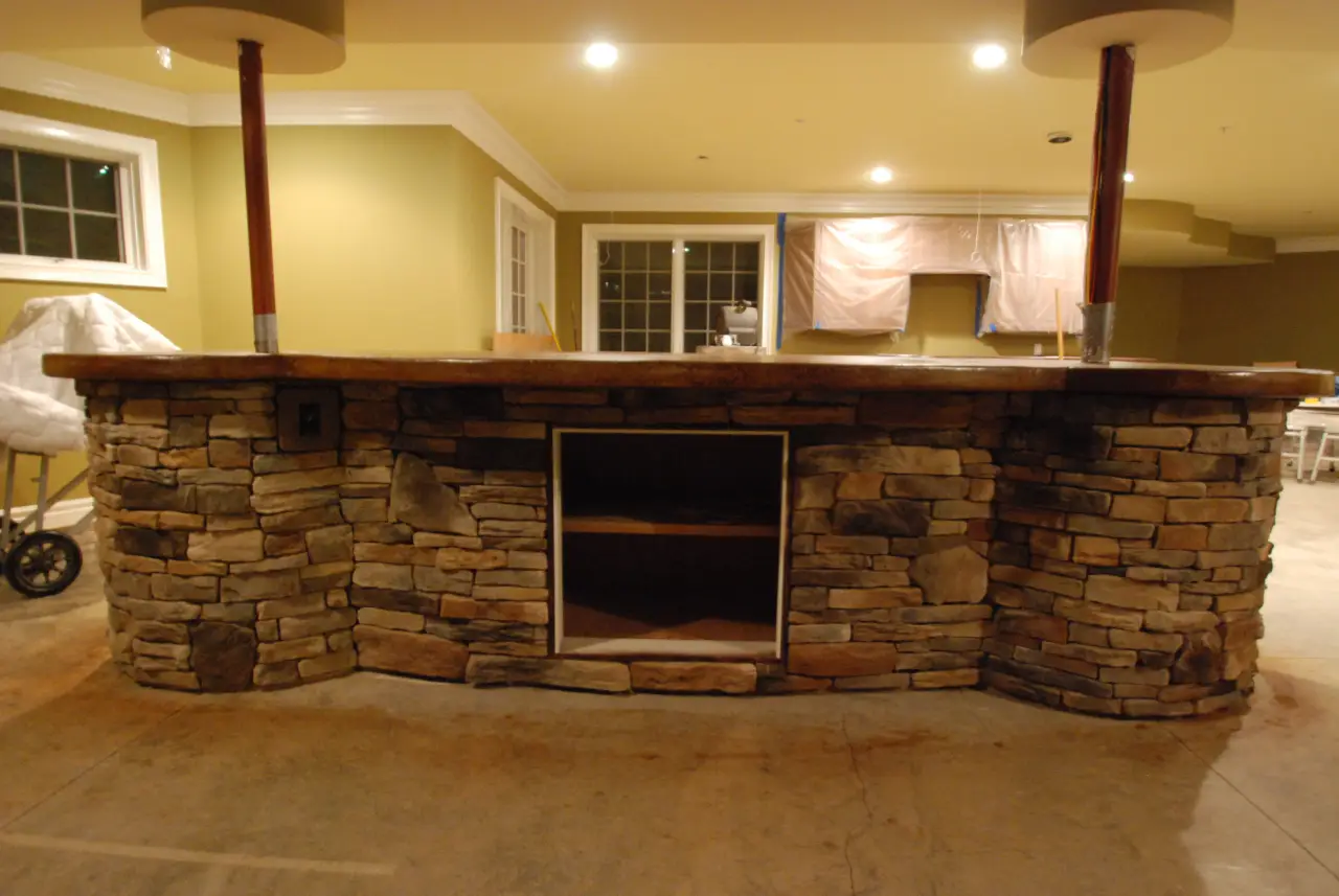 A stone bar with a wooden countertop, featuring two support poles, set in a partially finished room.