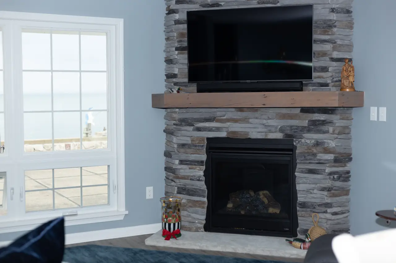 A cozy living room featuring a stone fireplace, a wooden mantel, and a wall-mounted TV, with large windows offering a view outside.