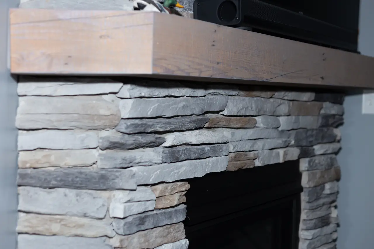 A stone fireplace with a wooden mantel, featuring a mix of light and dark stones, set against a gray wall.