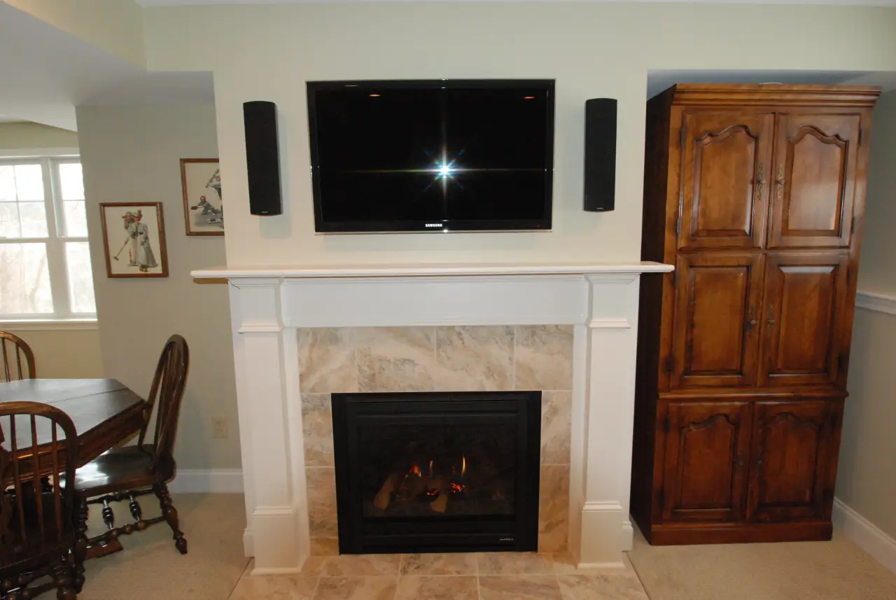 A cozy living room featuring a fireplace with a TV mounted above, flanked by speakers, and a wooden cabinet nearby.