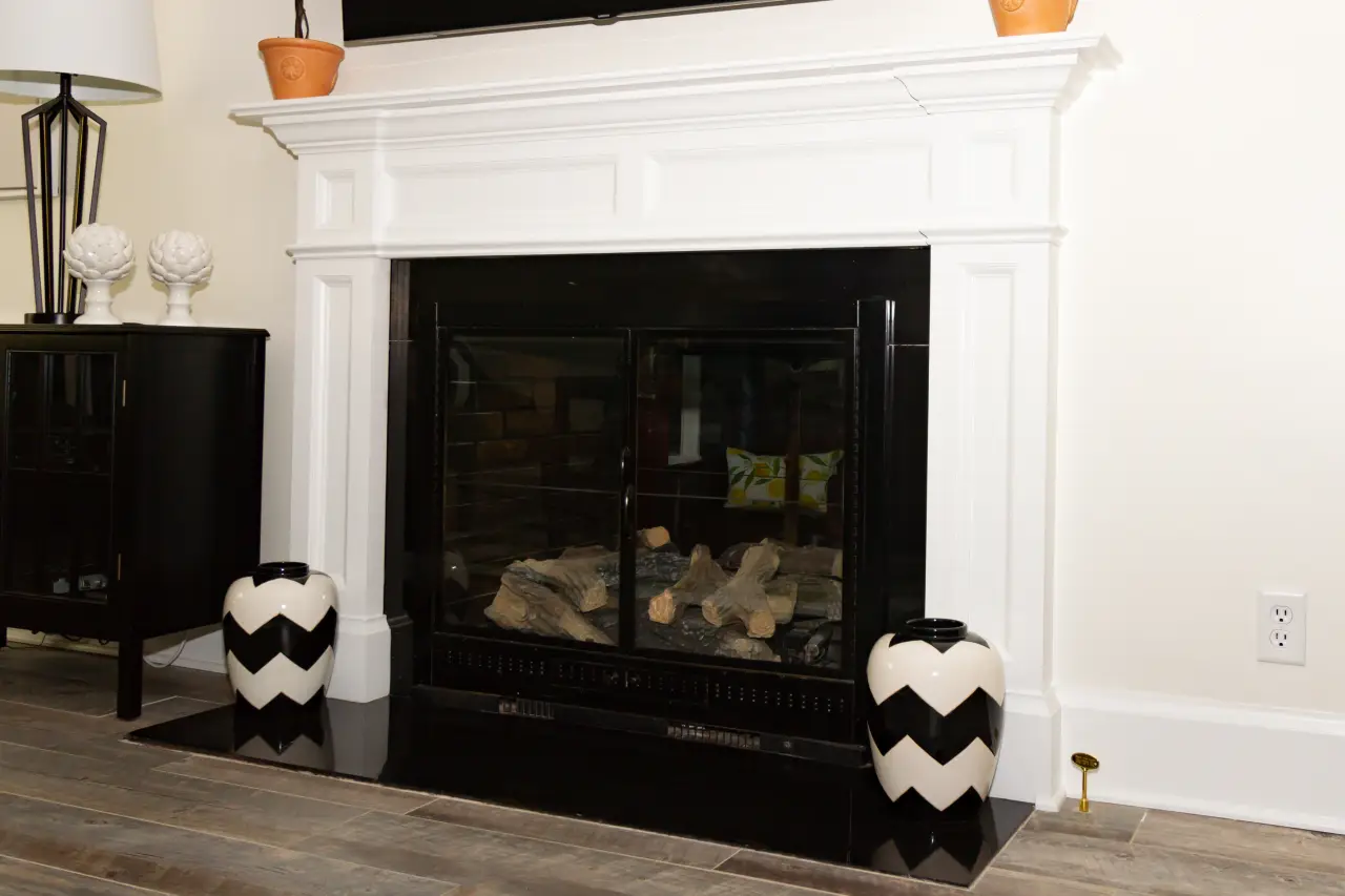 A modern fireplace with a white mantel, black glass doors, and two black and white chevron vases on either side.