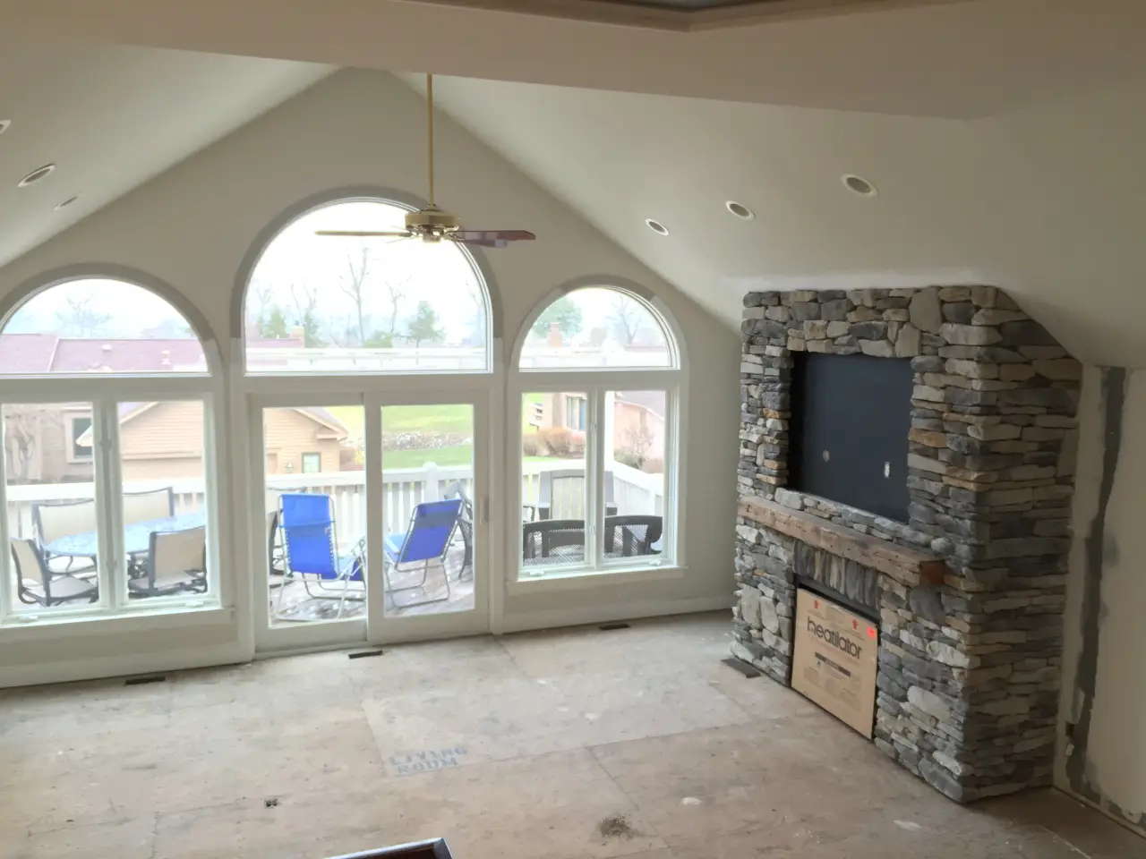 A spacious, unfinished living room with a stone fireplace, large arched windows, and a view of outdoor seating.