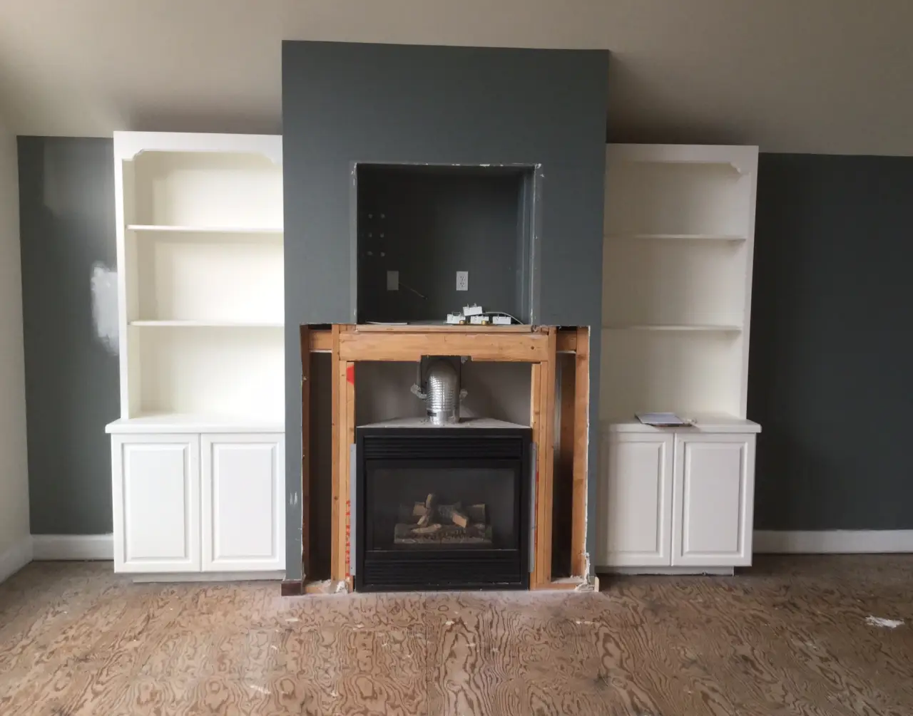 A room with unfinished walls, featuring a central fireplace framed by two white bookshelves. The floor is wooden and bare.