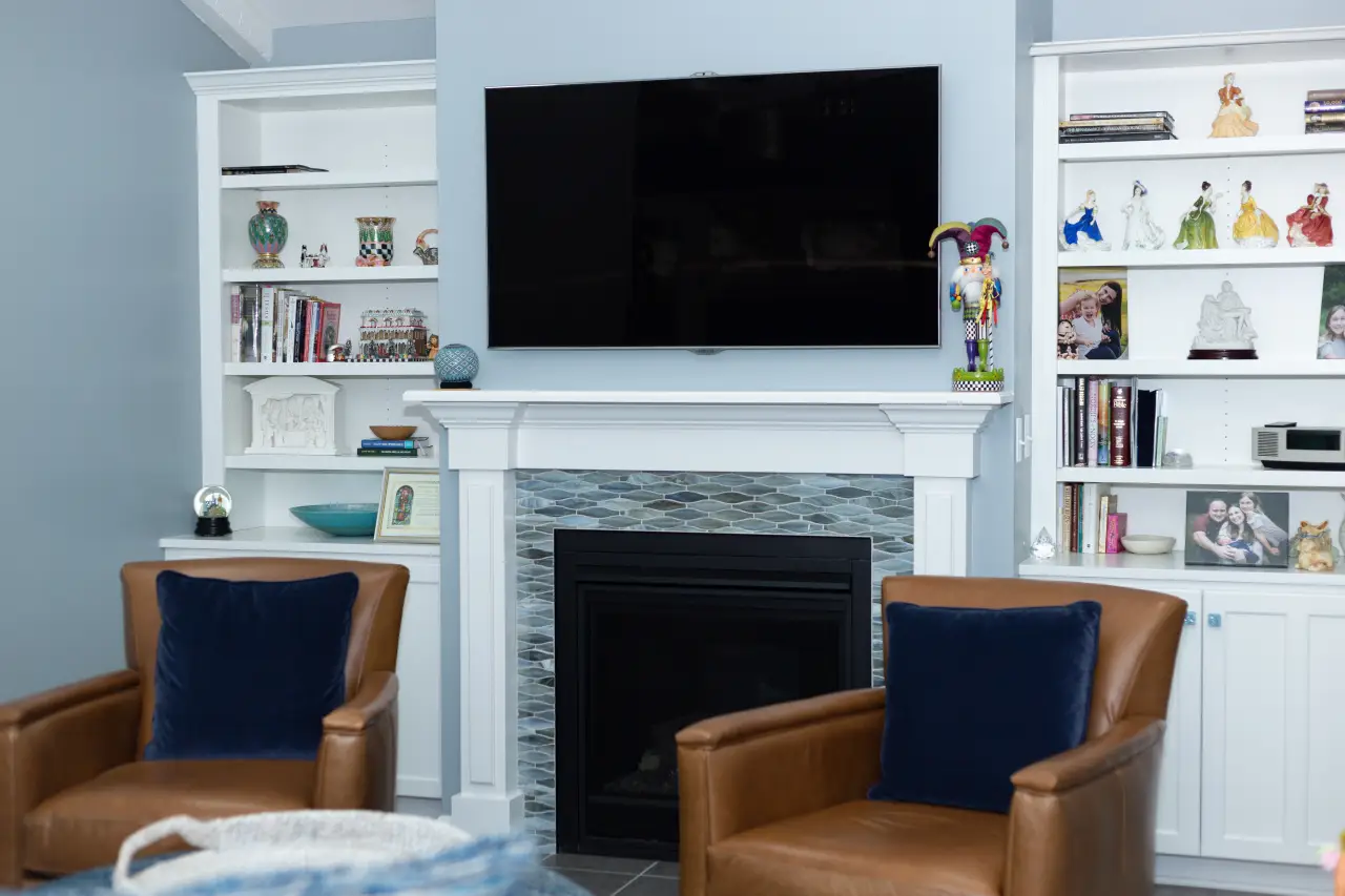 A cozy living room featuring a mounted TV above a fireplace, flanked by white bookshelves filled with decor and photos. Two brown armchairs are visible.