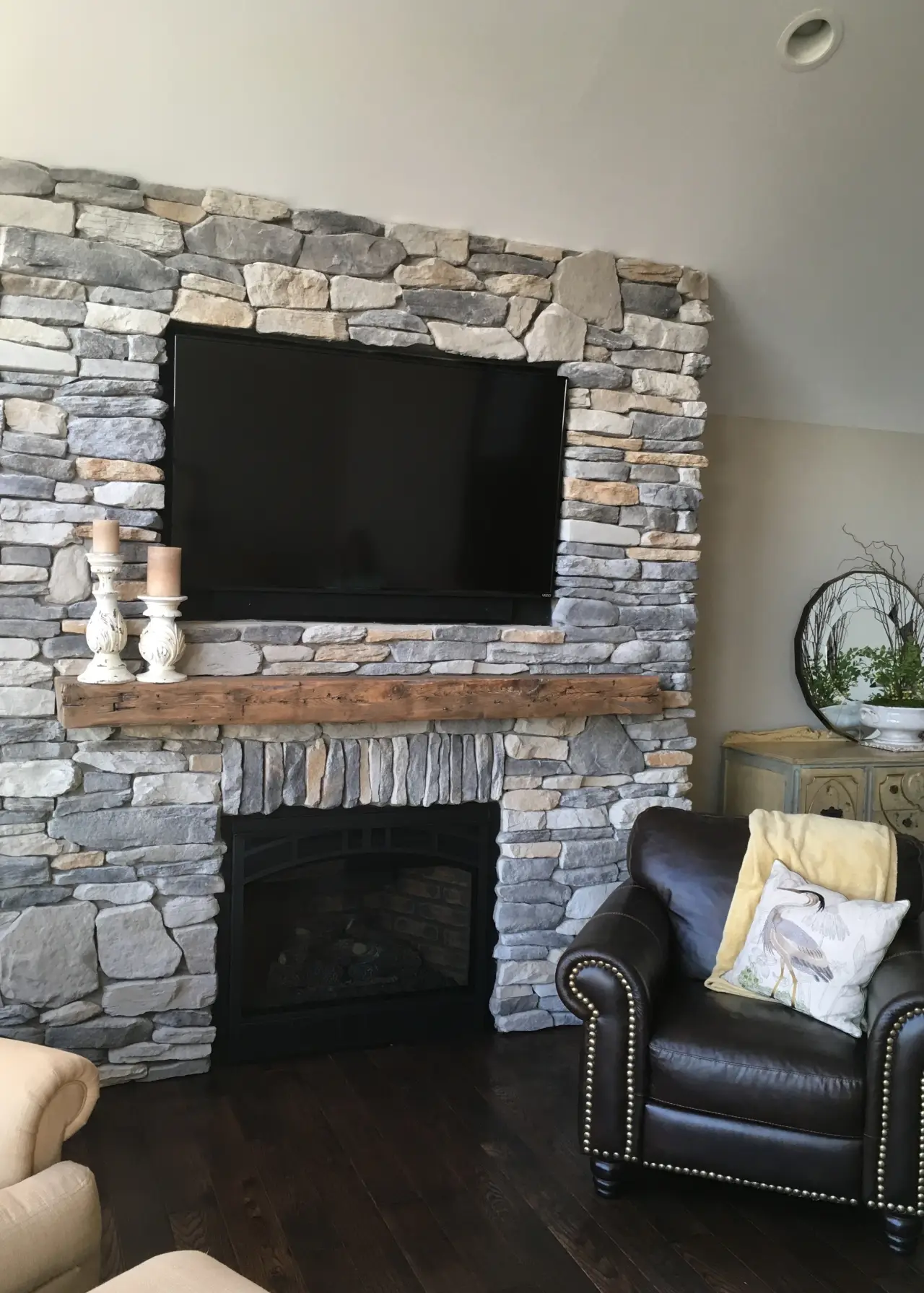 A cozy living room featuring a stone fireplace, a mounted TV, wooden mantel, and a leather armchair with decorative pillows.