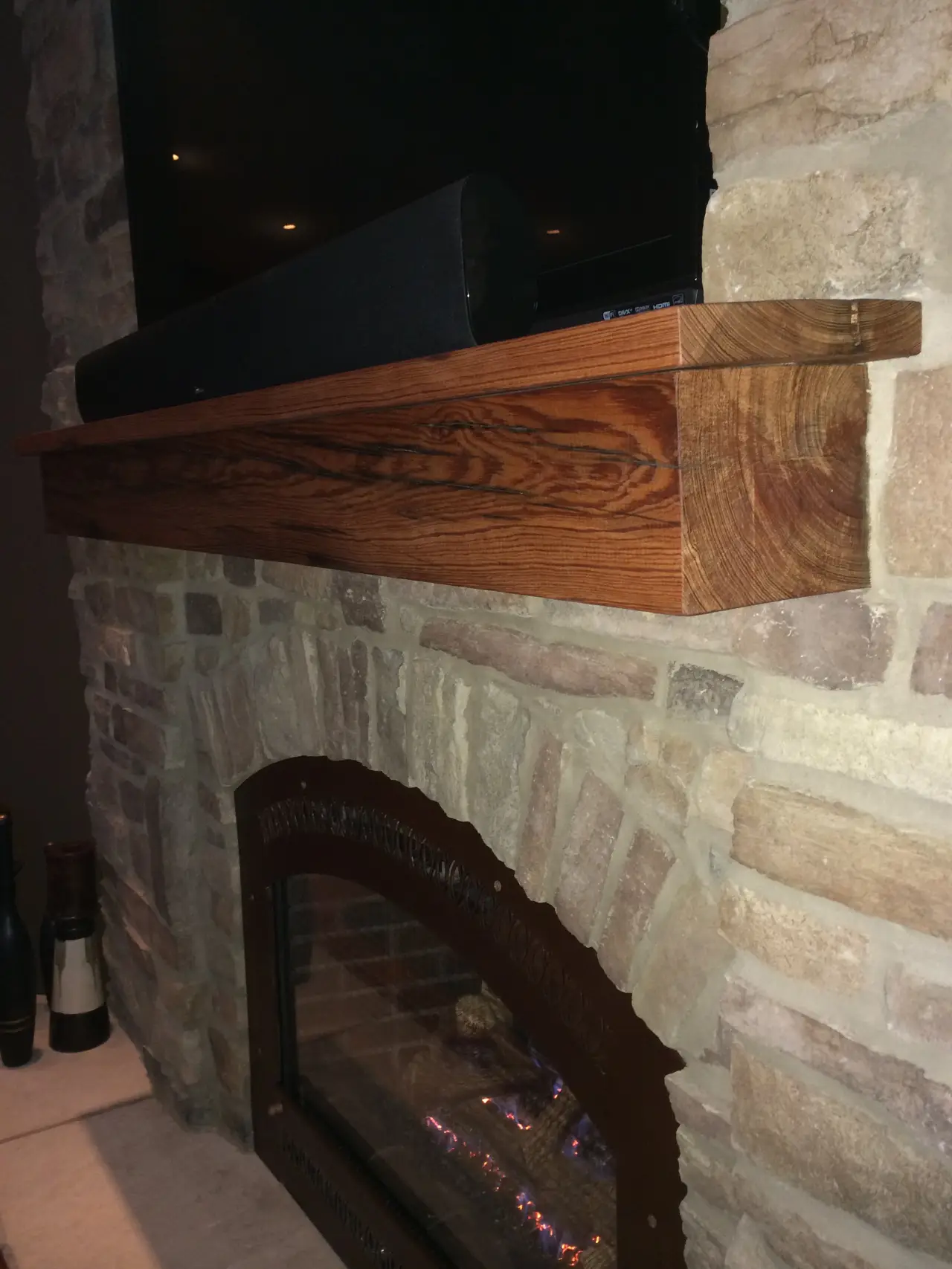 A wooden mantel rests above a stone fireplace, with a soundbar placed on top. The setting is cozy and modern.