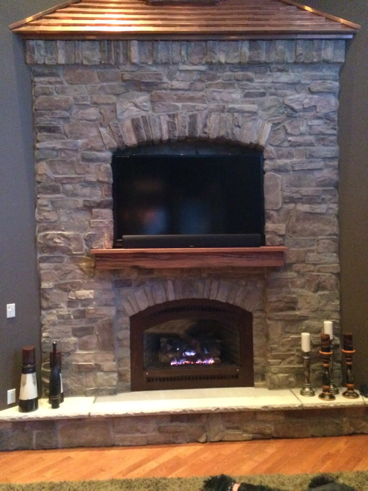 A stone fireplace with a wooden mantel, featuring a mounted TV above and a cozy fire below. Decorated with candles and vases.