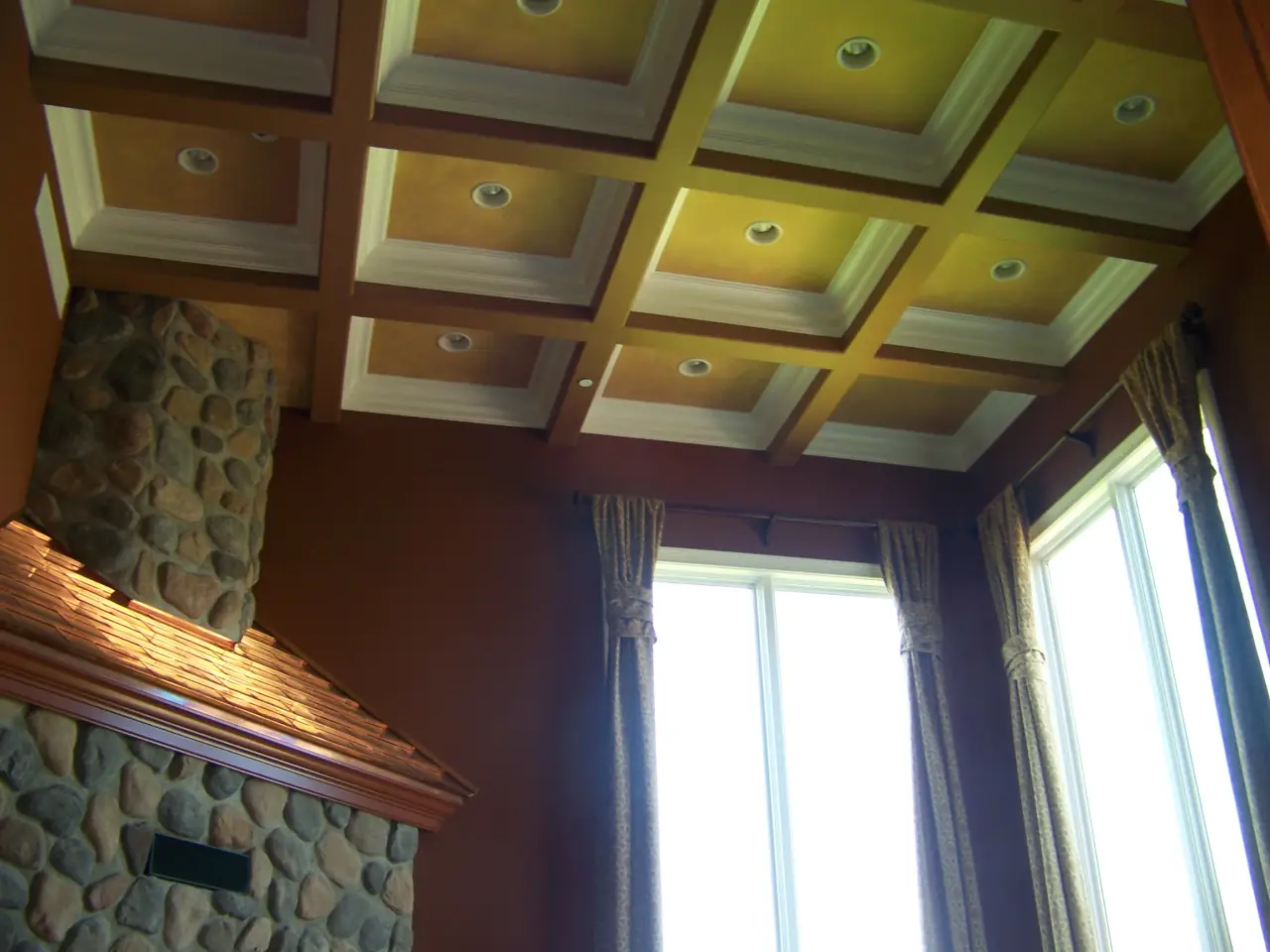 A room with a coffered ceiling, stone fireplace, and large windows dressed with curtains.