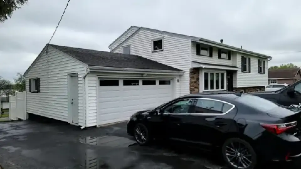 A black car is parked in front of a white two-story house with an attached garage.