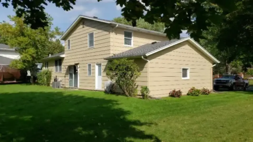 Tan two-story house with a sloped roof and green lawn, surrounded by trees and shrubs.