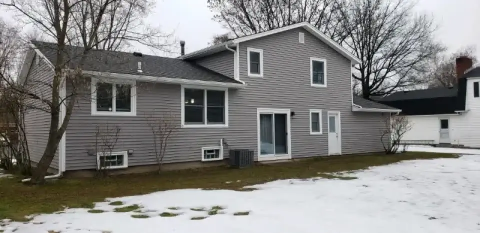 Gray two-story house with bare trees, some snow patches, and a cloudy sky in the background.