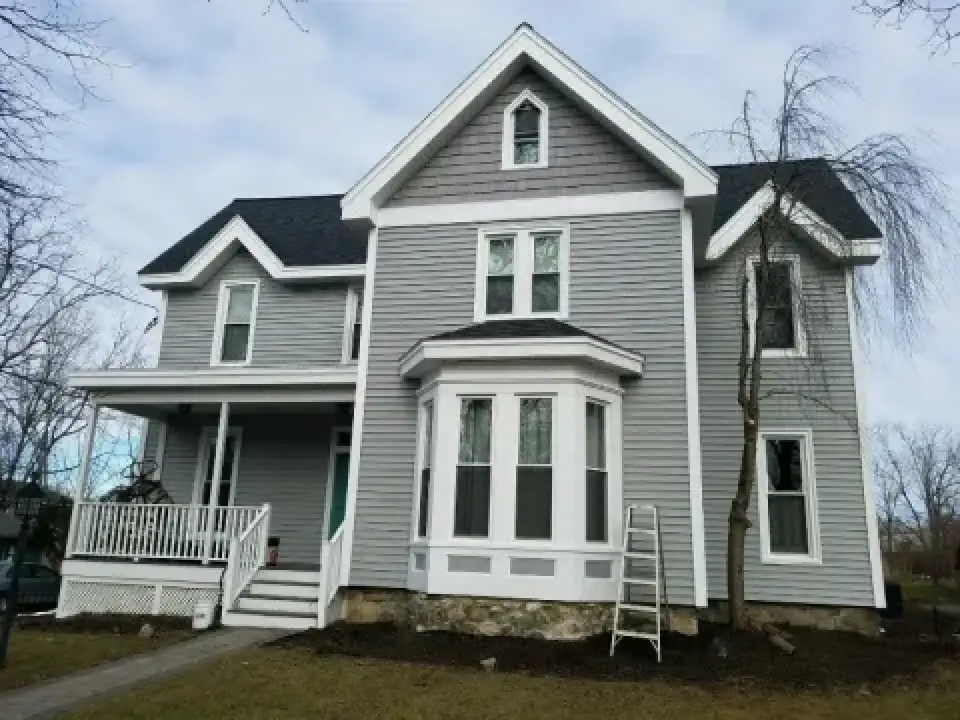 Two-story gray house with white trim, front porch, and a ladder leaning against the wall.