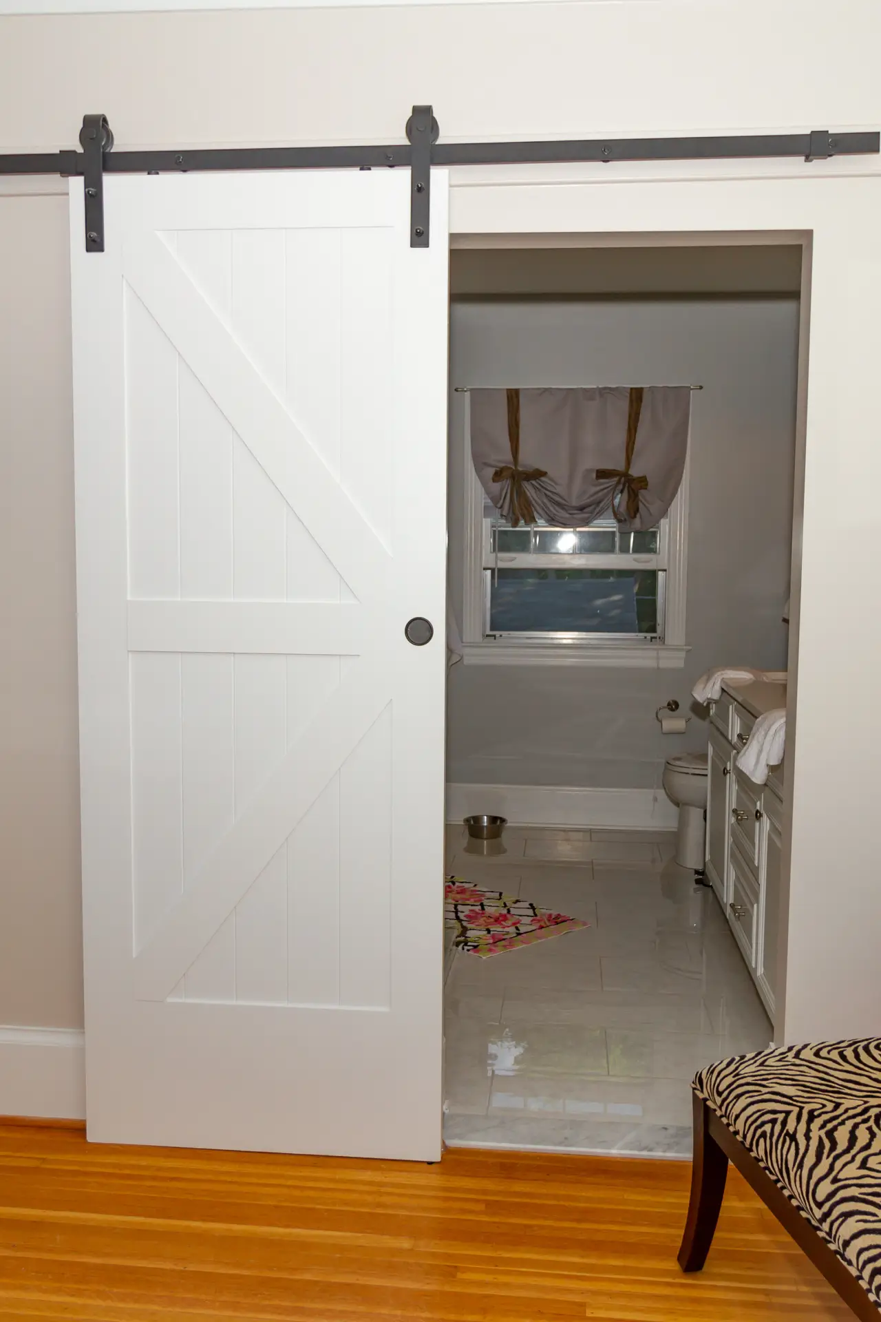 A white sliding barn door partially opens to reveal a bathroom with a window, towels, and a colorful rug on the floor.