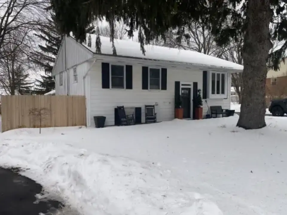 White house with black shutters and porch chairs, surrounded by snow and bare trees in winter.