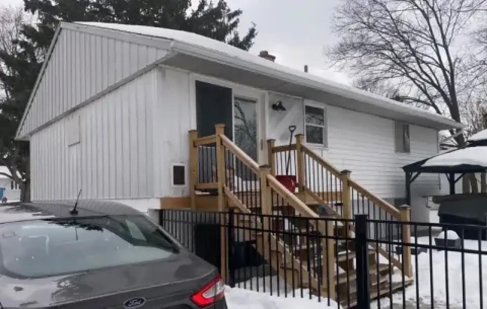 Single-story white house with wooden steps, snow on the ground, and a parked gray car nearby.