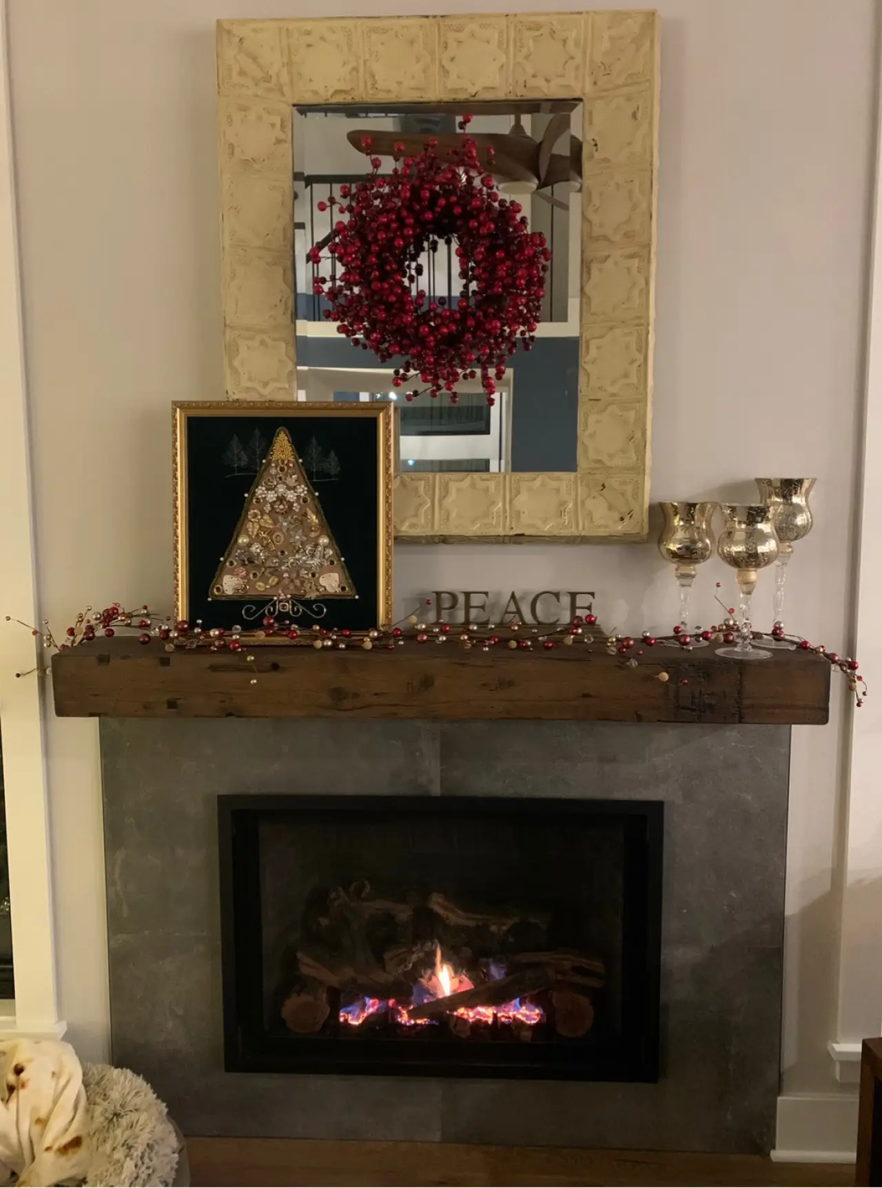 A cozy fireplace with a wooden mantel, decorated with a wreath, a framed tree art piece, and silver cups, featuring a glowing fire below.