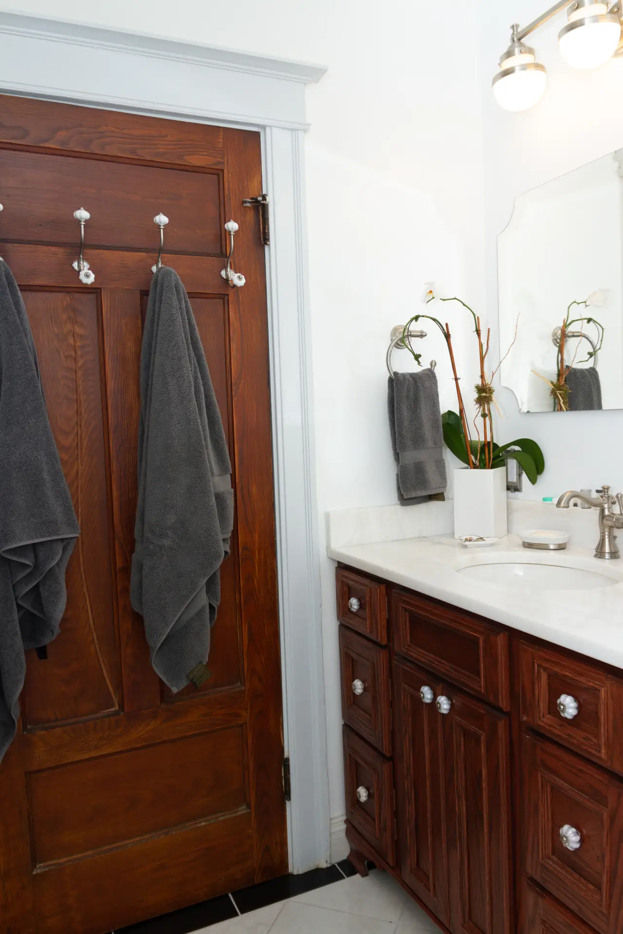 A modern bathroom featuring a wooden door, gray towels, a marble countertop, and a mirror with stylish lighting.