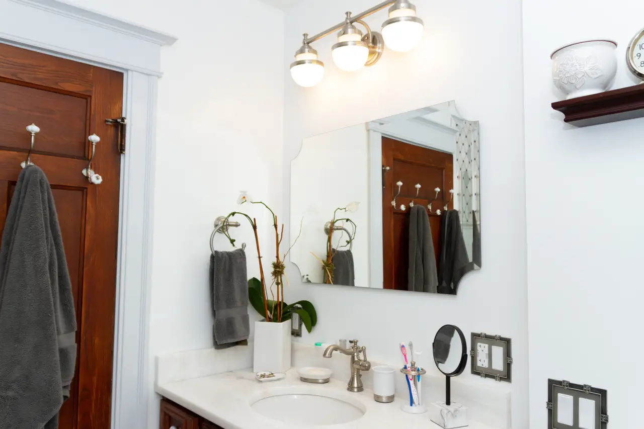 A modern bathroom featuring a mirror, light fixtures, a sink with toiletries, and towels hanging on hooks.