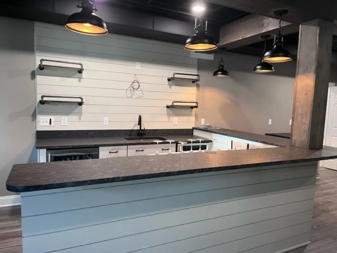 A modern kitchen with a dark countertop, light wood paneling, and pendant lights. Features open shelving and a sink area.