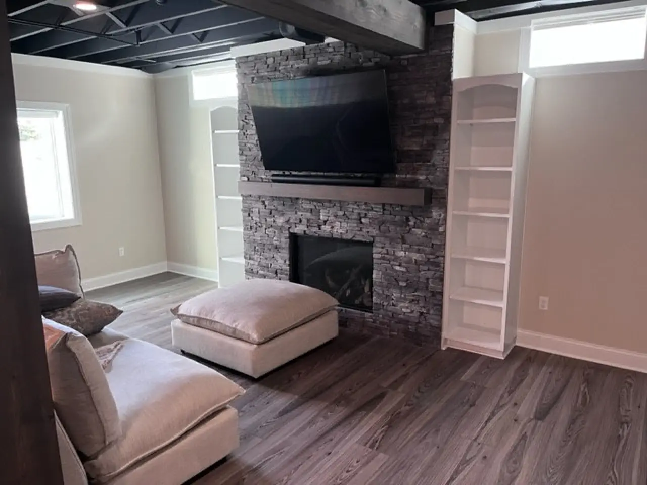 A modern living room featuring a stone fireplace, a mounted TV, and two white bookshelves, with cozy seating and wooden flooring.