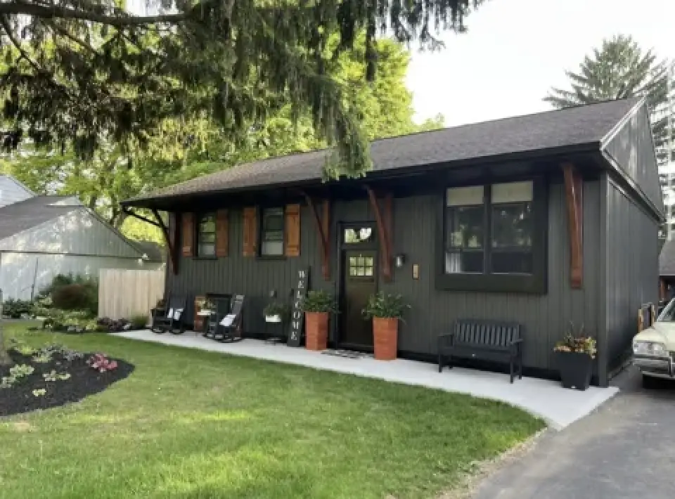 A dark green ranch-style house with a porch, potted plants, and outdoor seating.
