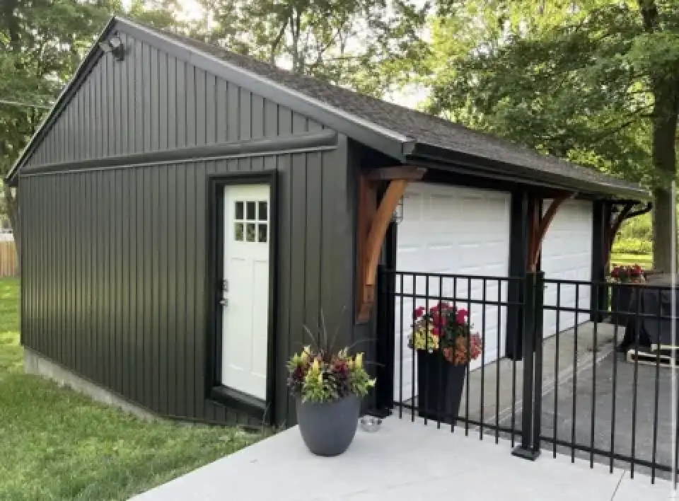 Black-sided garage with white door, potted plants, and a black fence in a green yard.