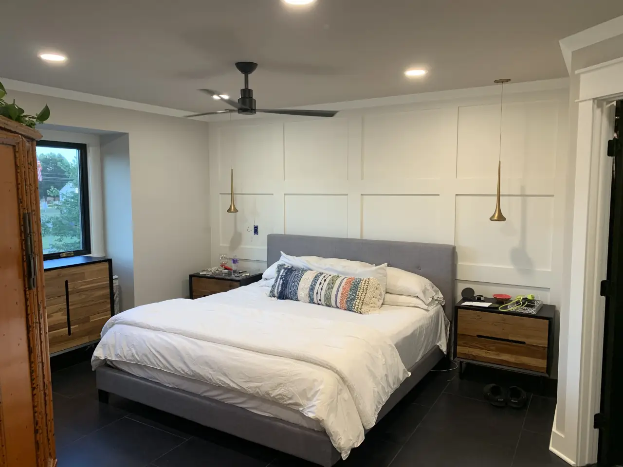 A modern bedroom featuring a gray bed with white bedding, wooden nightstands, pendant lights, and a large window.