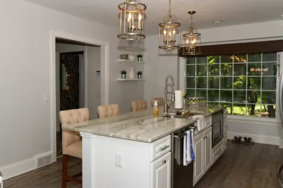 Modern kitchen with marble island, pendant lights, bar stools, and large window overlooking greenery.