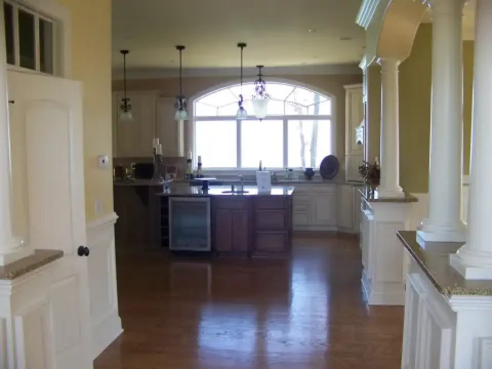 Spacious kitchen with wooden floors, large window, island, white cabinets, and hanging lights.