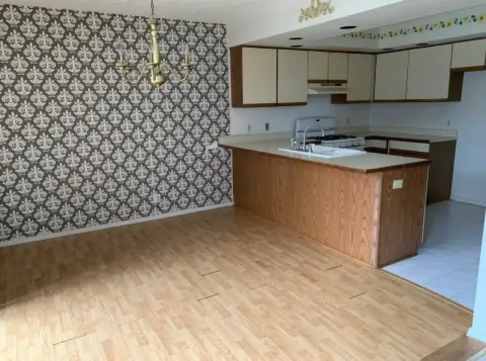 Kitchen with wood cabinets, patterned wallpaper, chandelier, and light wood flooring in dining area.