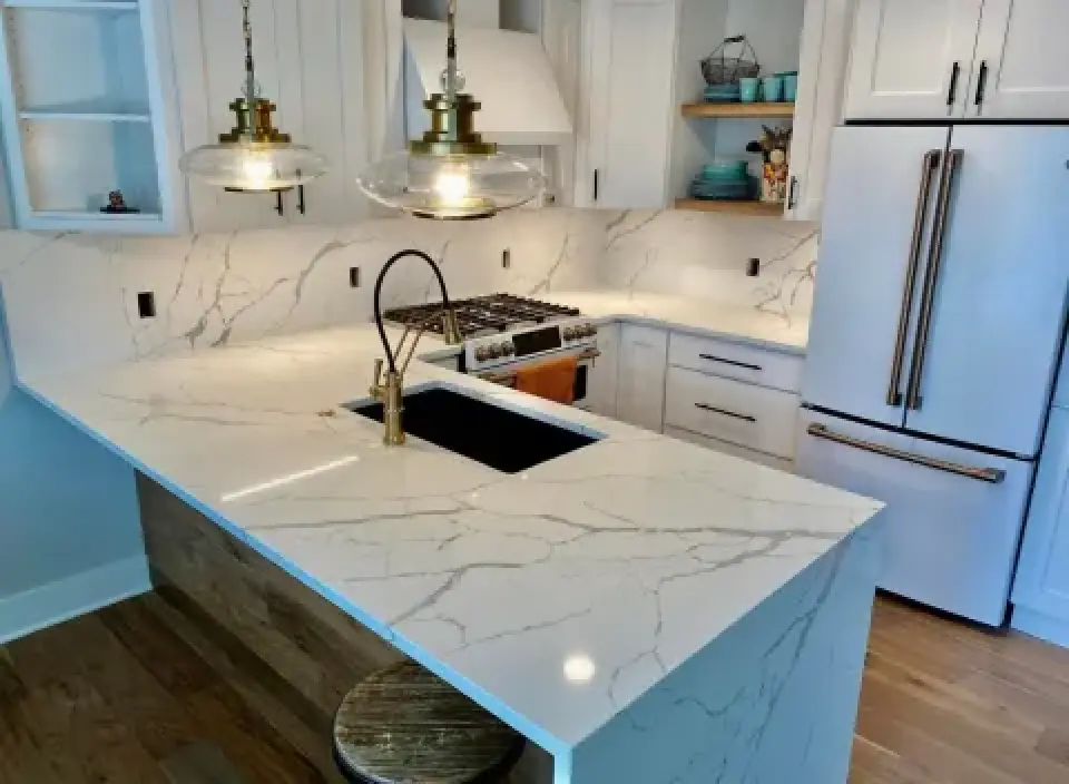 Modern kitchen with white marble countertops, gold faucet, pendant lights, and stainless fridge.