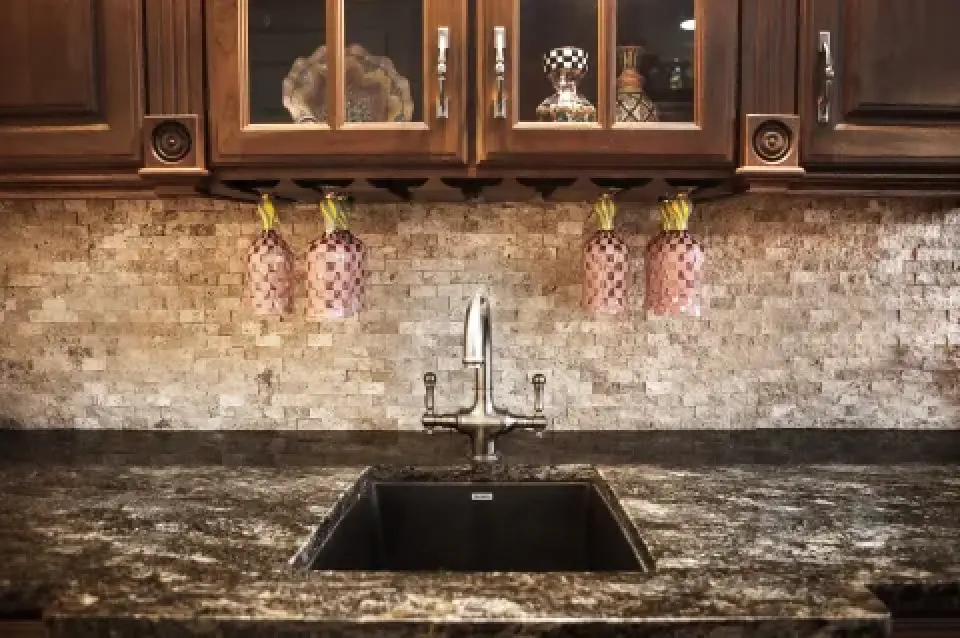 Kitchen sink with dark marble countertop, wooden cabinets, and hanging decorative wine glasses.