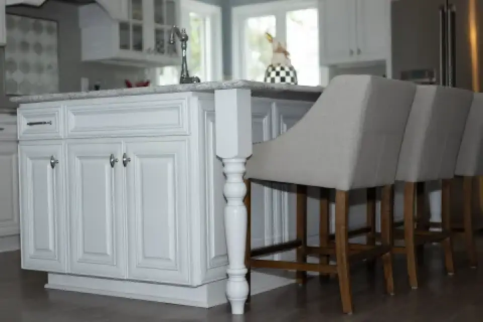 White kitchen island with carved leg and two beige barstools on a wooden floor.