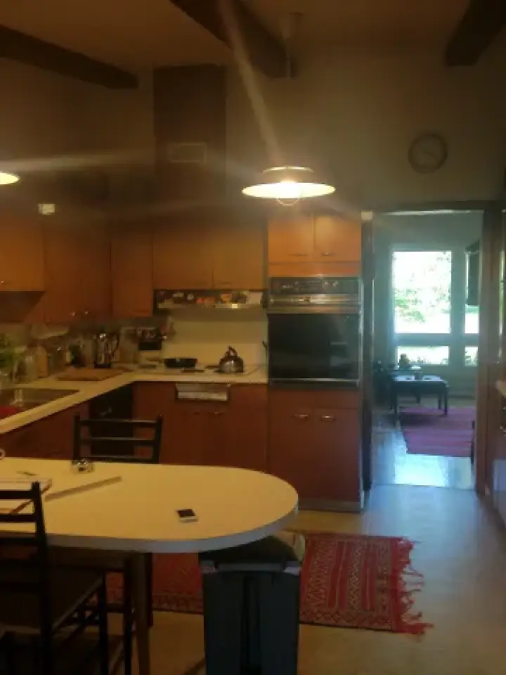 A cozy kitchen with wooden cabinets, a round table, and rugs, leading to a sunlit room.
