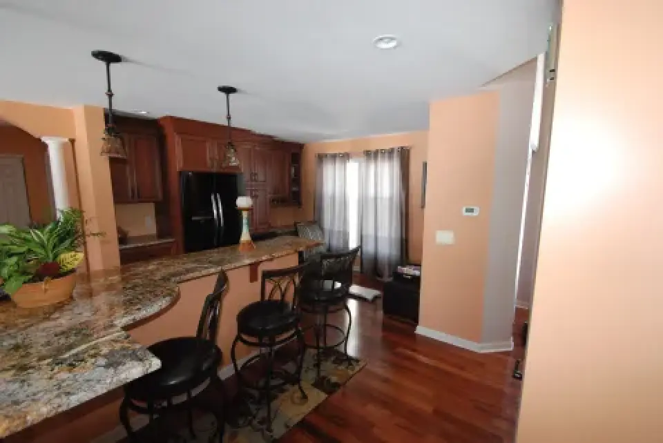 Kitchen with granite island, bar stools, wooden floors, cabinets, and sunlight through curtains.