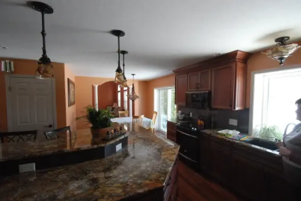A kitchen with brown cabinets, marble counters, and a person standing near a window.