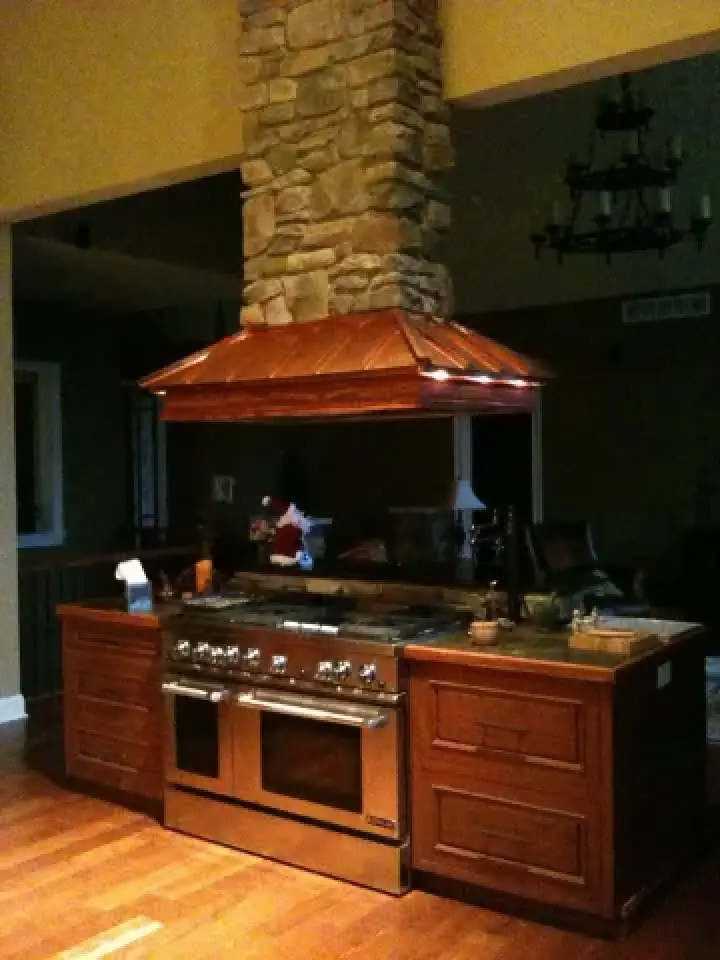 Kitchen island with a stove, copper range hood, stone chimney, and wooden cabinets.