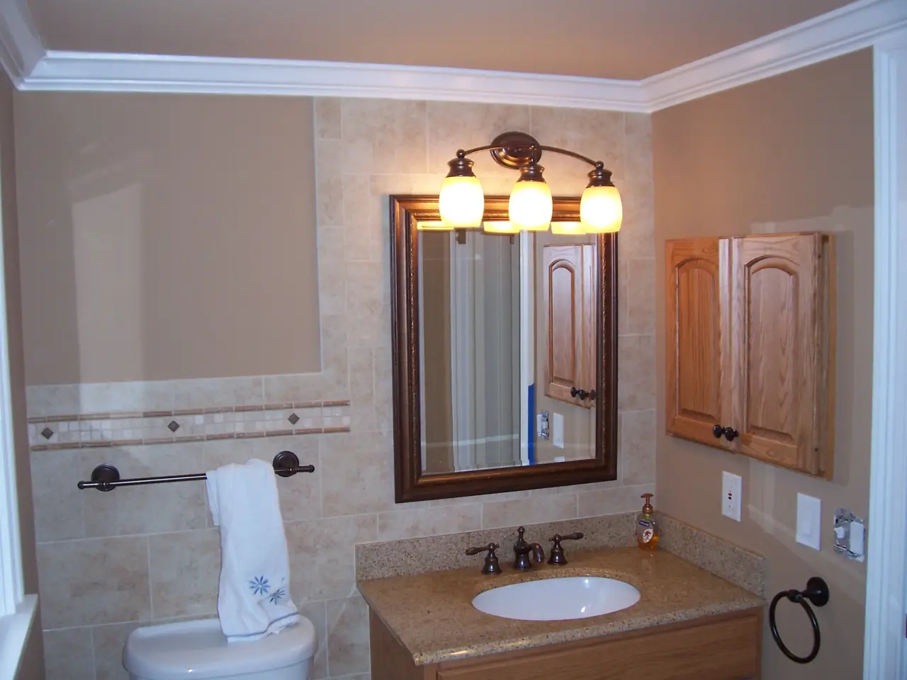 A modern bathroom featuring a wooden cabinet, a round sink, a mirror with lights, and beige tiled walls.