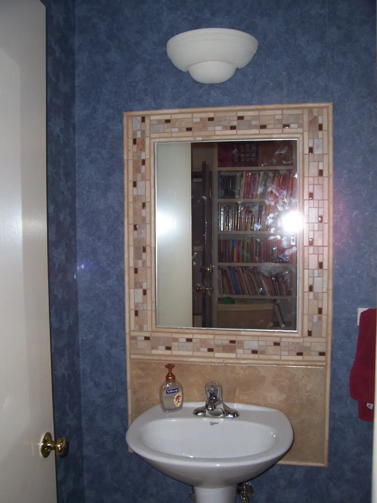 A small bathroom featuring a wall-mounted mirror, sink, and a towel. Bookshelves are visible in the reflection.