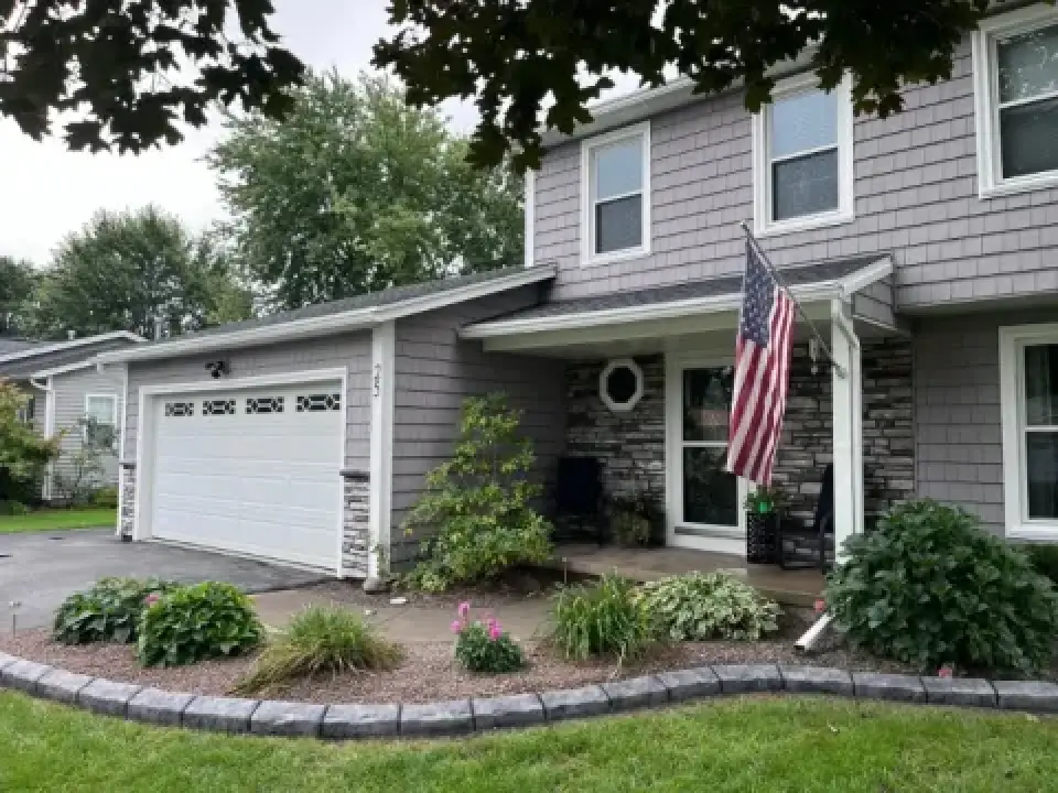 Gray house with American flag, front porch, garden beds, and two-car garage on a cloudy day.