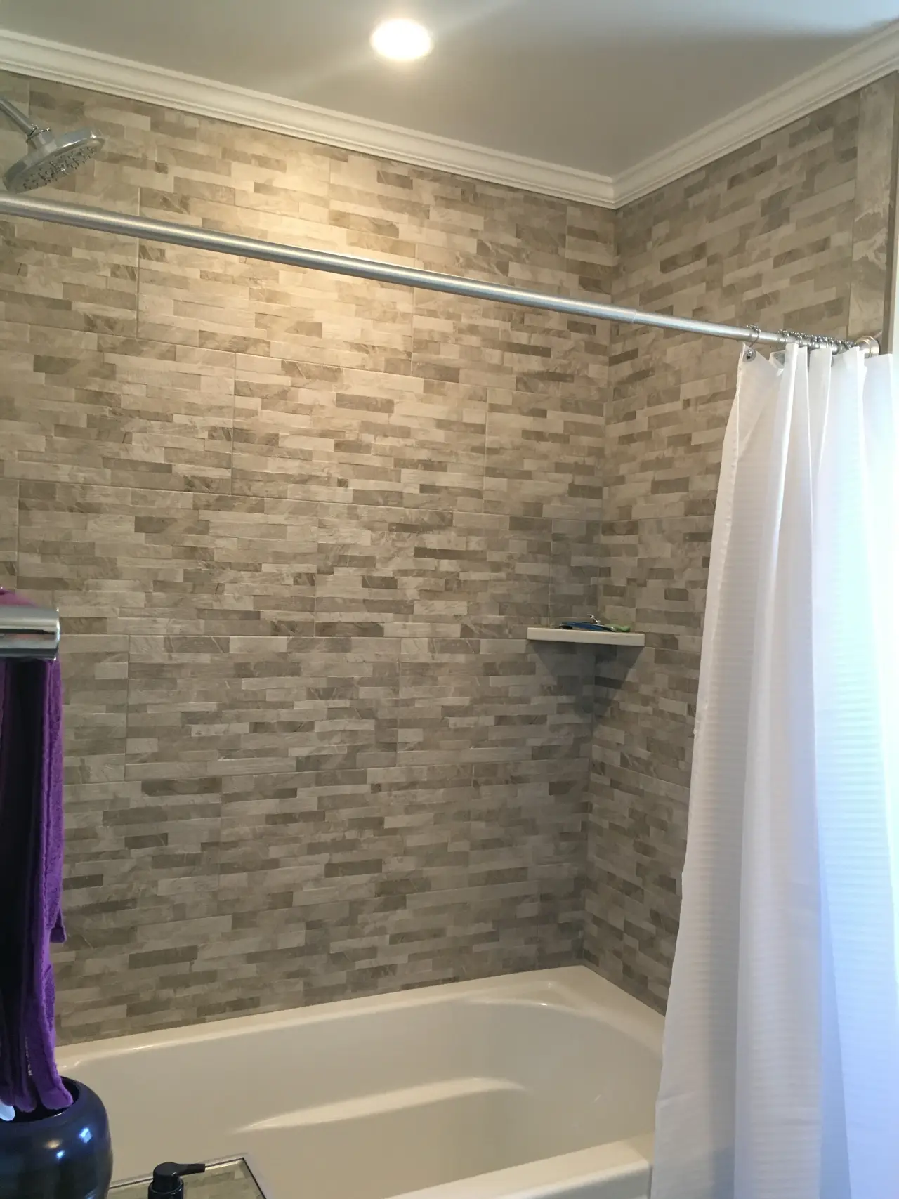 A modern bathroom with gray stone tile walls, a white shower curtain, and a bathtub. A small shelf is visible on the wall.